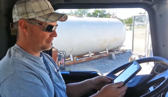 A propane technician checks a rugged tablet in the cab, with a stationary propane tank visible through the windshield — verifying records before the job.